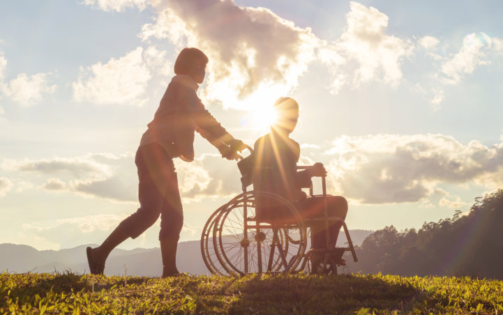 a man in a wheelchair strolling along the outdoors with assistance